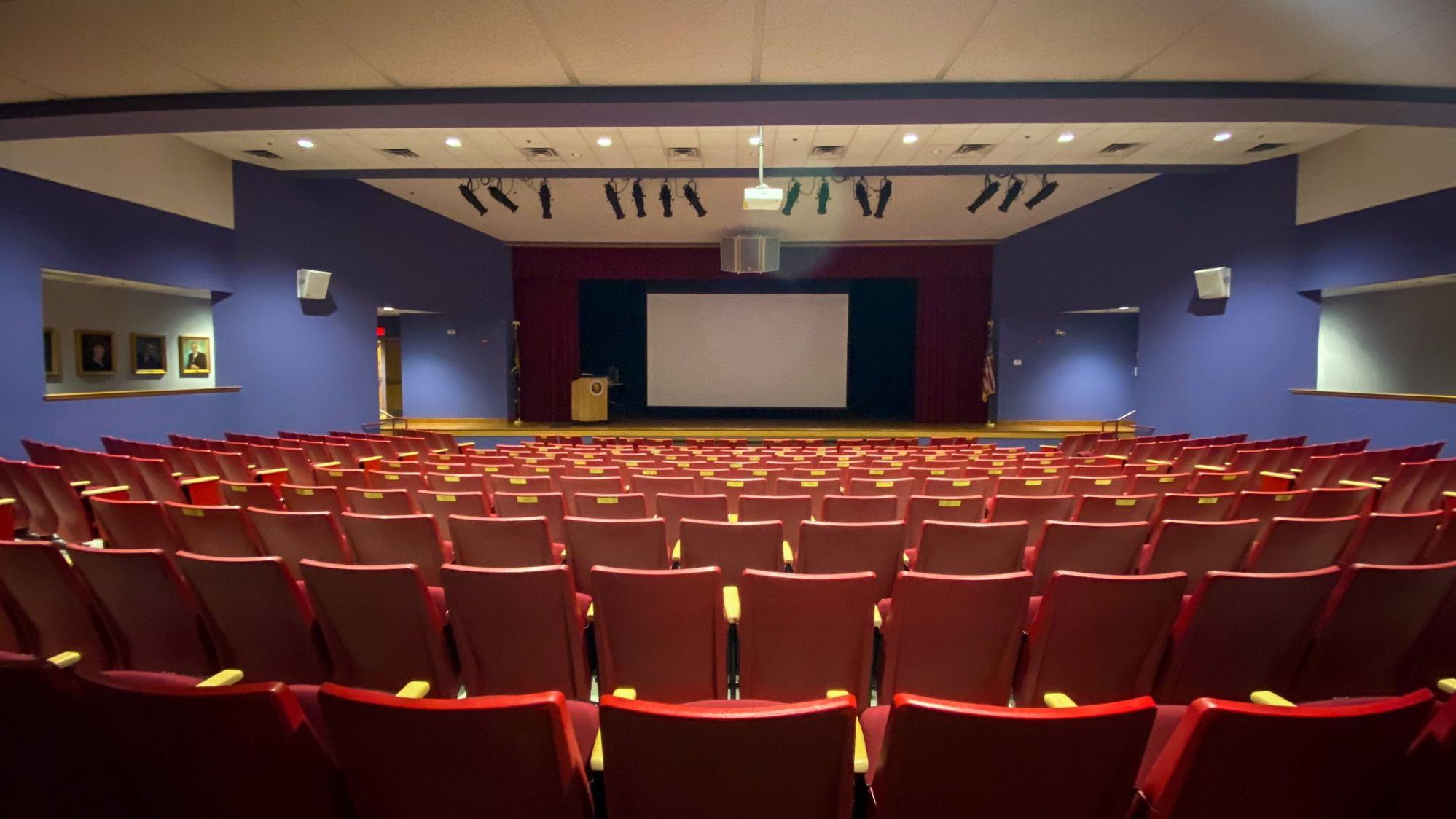 Auditorium stage with projector screen down; many red auditorium seats are in the foreground.